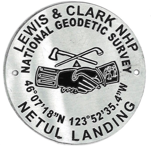 A hiking stick medallion depicting Netul Landing's National Geodetic Survey marker. Text reads "Lewis & Clark NHP. National Geodetic Survey. 46 7' 18"N 123 52' 35.4" W Netul Landing" with two hands shaking and a crossed pipe and tomahawk.